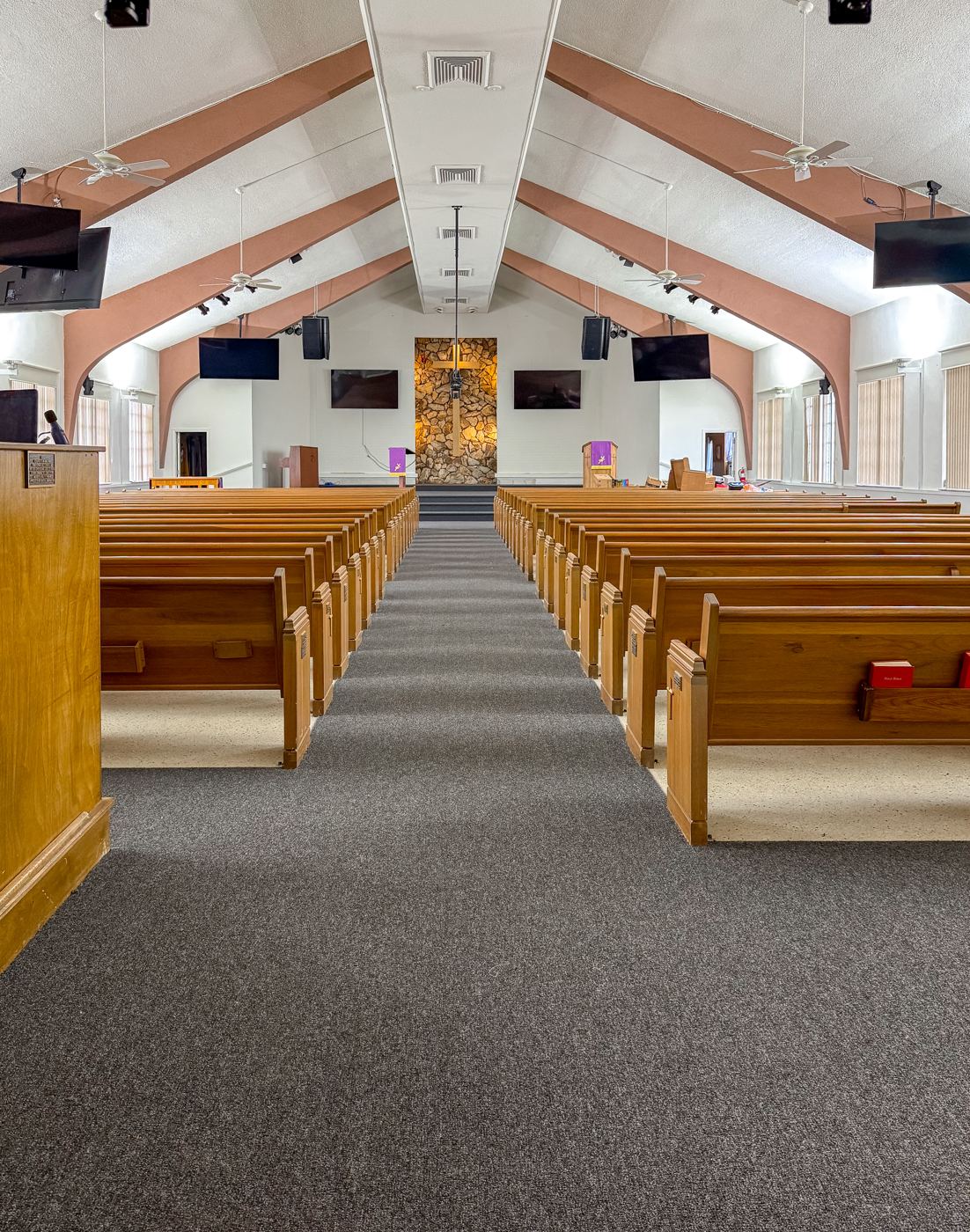 Finished worship hall with new gray carpet, refurbished wood pews, fresh paint, and the lit stone altar wall.