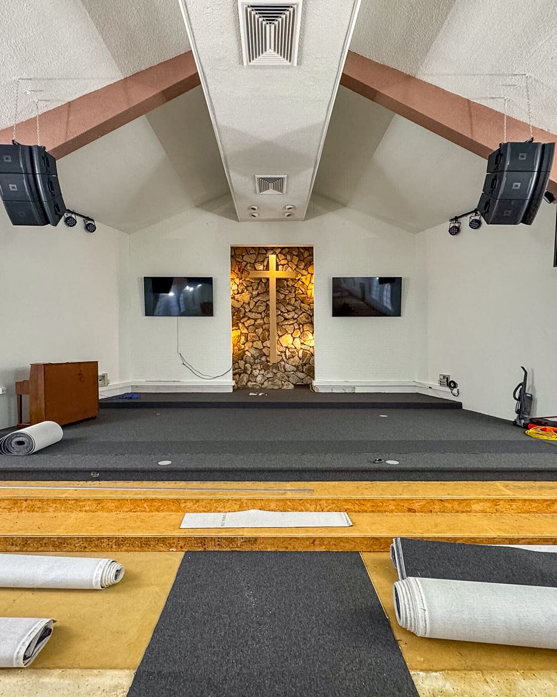 Church sanctuary mid-renovation: subfloor exposed, new carpet rolled and staged, lit stone altar wall and cross at the far end.