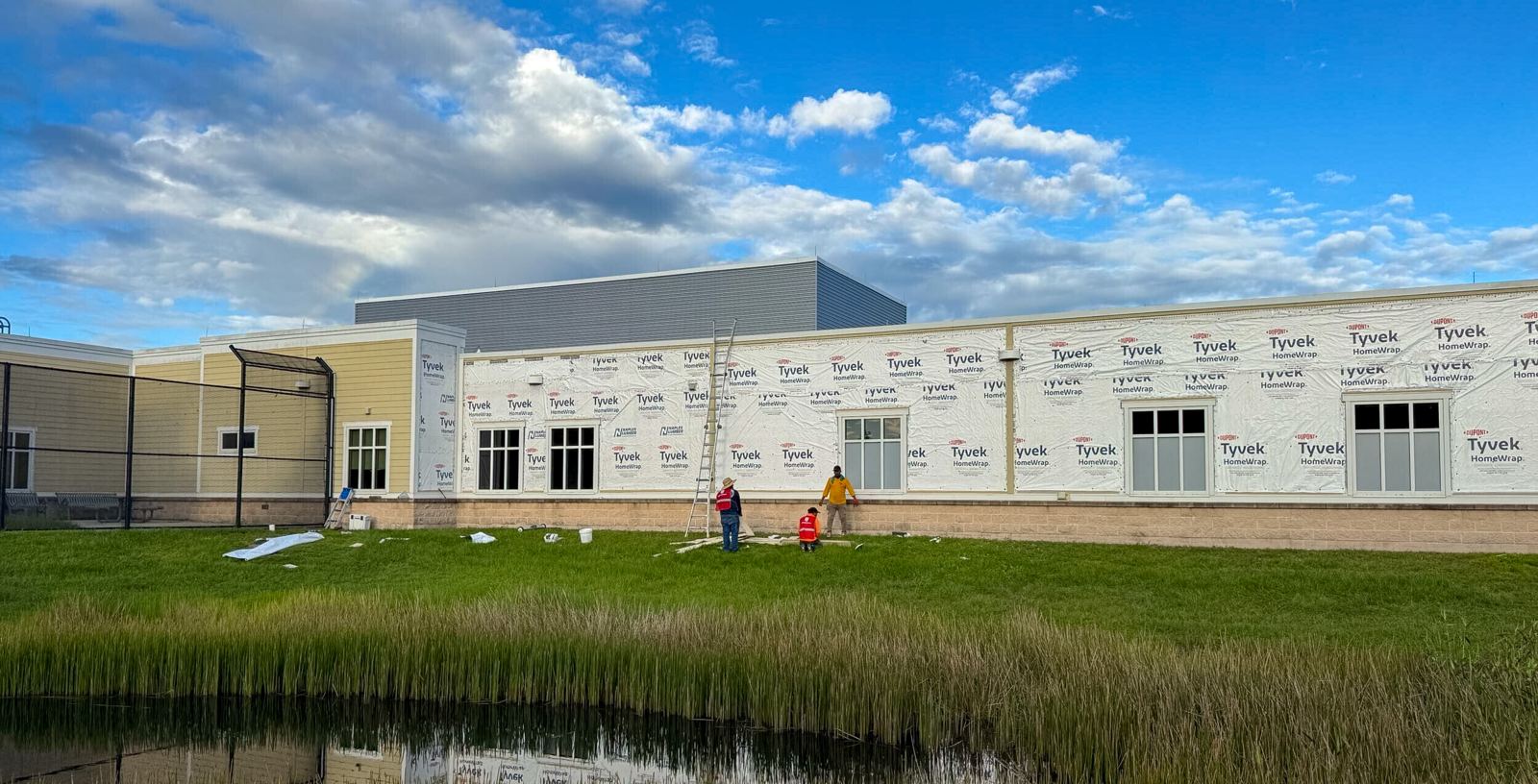 North Tampa Behavioral Health Hospital wrapped in new weather barrier during exterior restoration, crew at work, blue sky reflected in the pond in front.