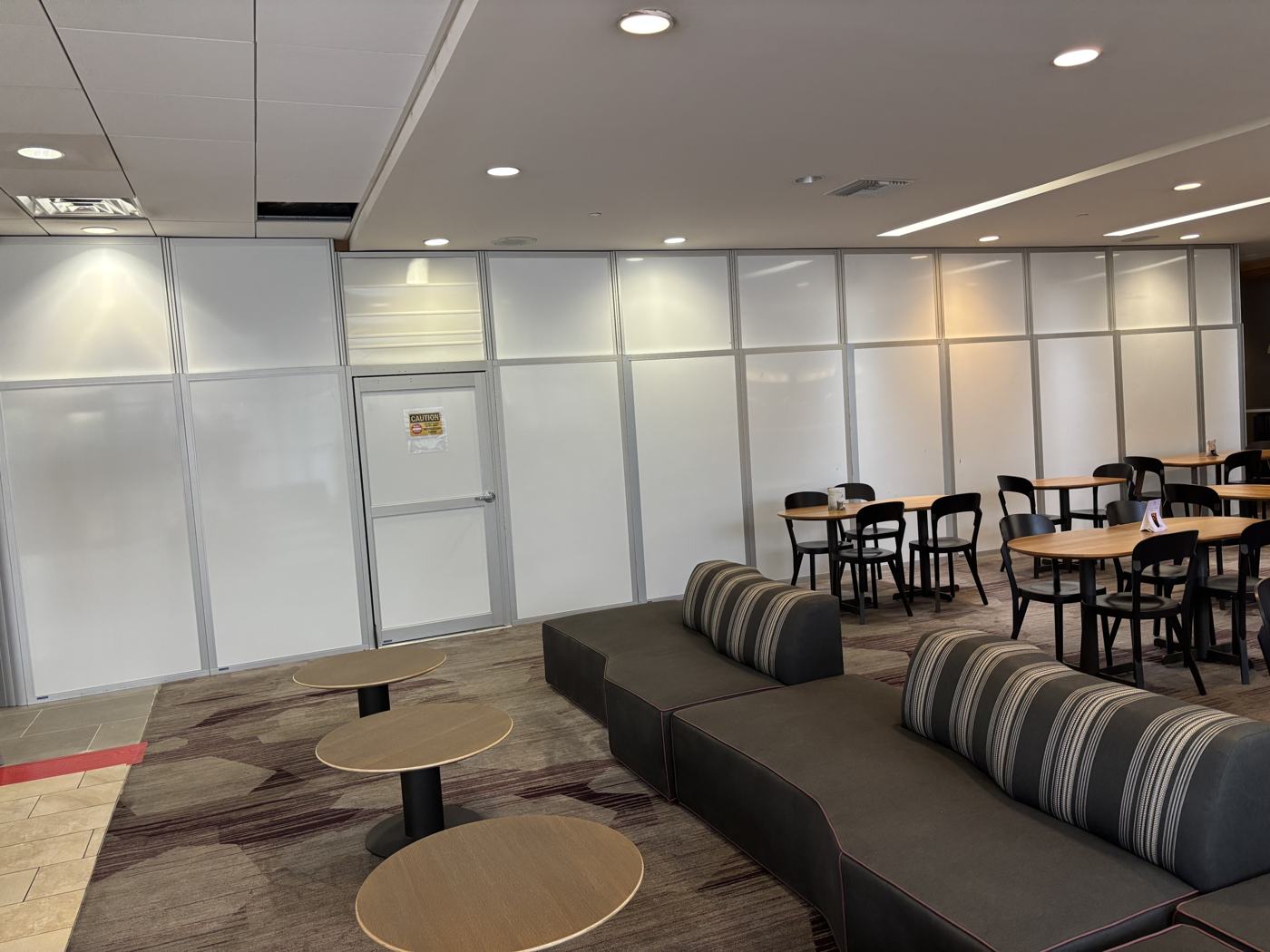 Hotel lobby in use: guests' seating and dining tables set up directly against a clean modular containment wall with a signed access door.