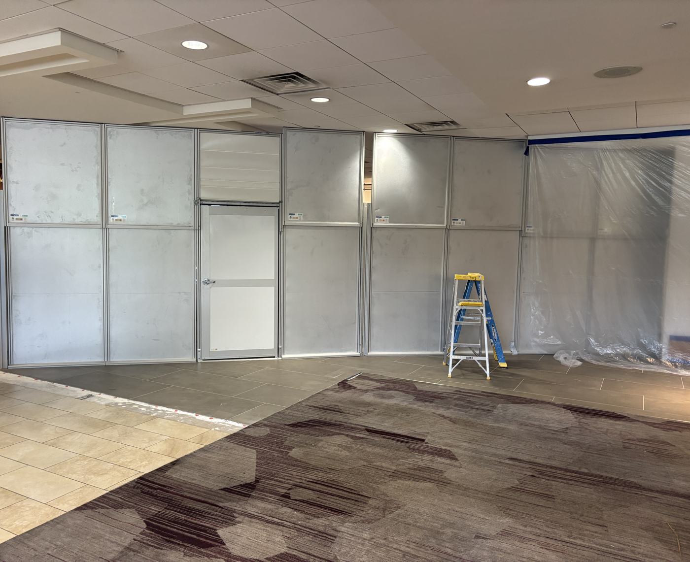Modular containment wall being assembled across a hotel lobby, with a lockable access door and plastic sheeting sealing the far end.